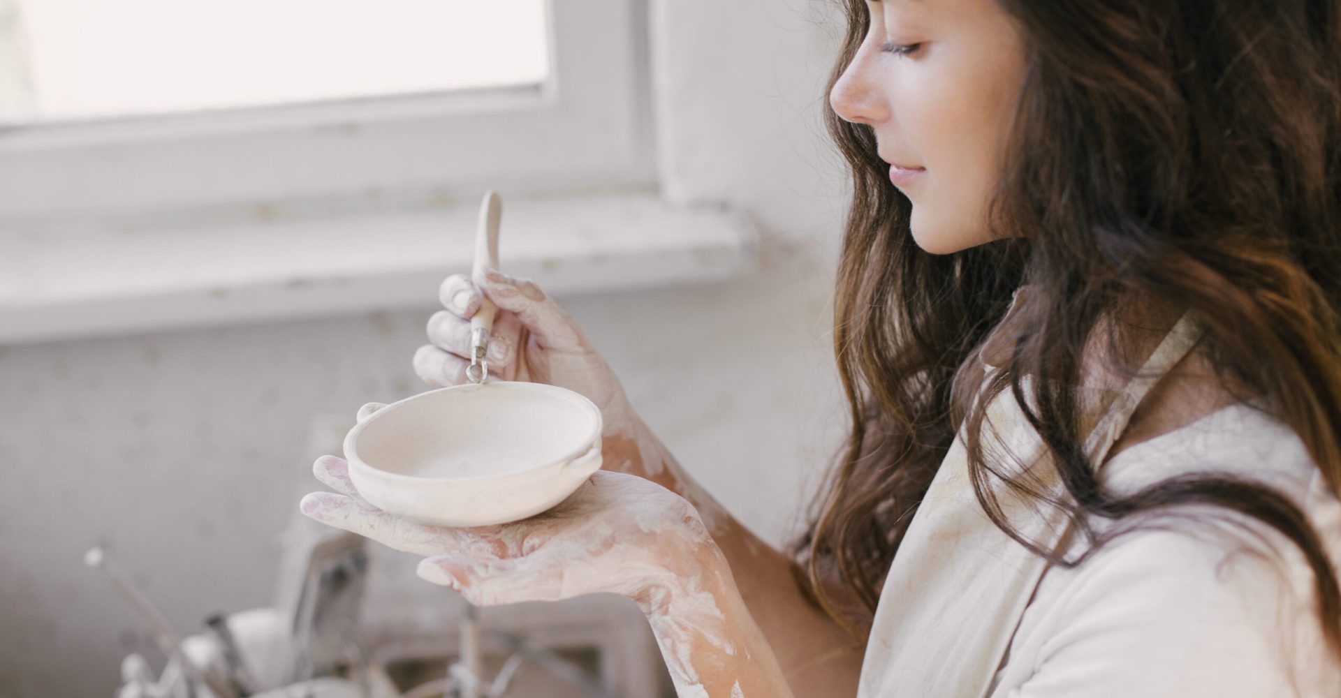 Young beautiful woman with long curly hair in white apron creating handmade ceramic bowl in a pottery. Creative workshop.